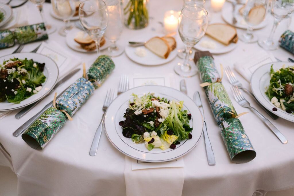 A round table set for a formal meal with plates of mixed green salad, wine glasses, candles, bread slices, and festive holiday crackers on white tablecloths.