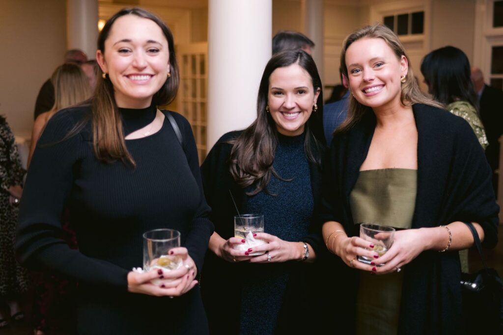 Three women dressed in semi-formal attire stand close together at an indoor event, each holding a drink and smiling at the camera. Other guests and white columns are visible in the background.
