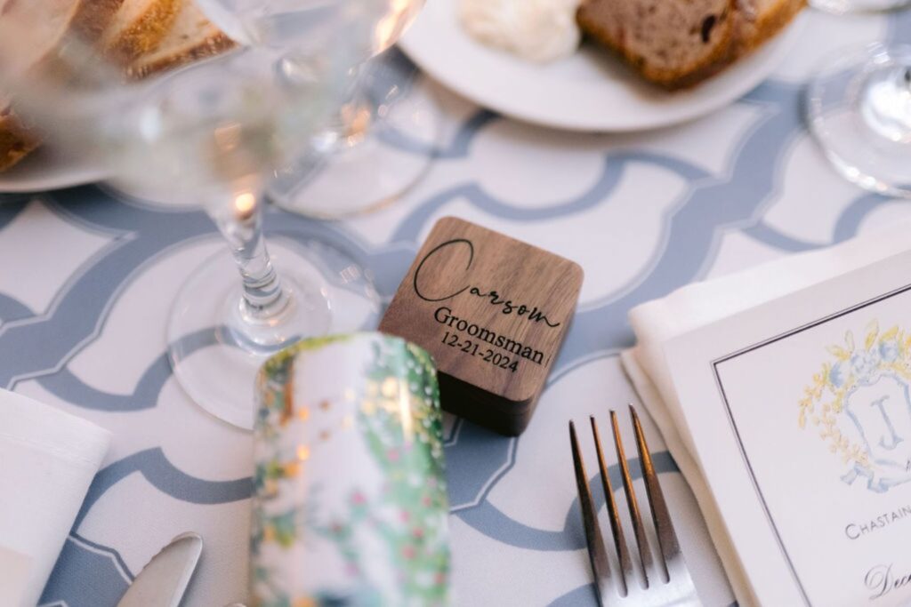 A close-up of a wedding table setting featuring a small wooden box labeled Carson, Groomsman, 12-21-2024, a menu, fork, knife, a glass, a plate with cake, and a party cracker on a patterned tablecloth.