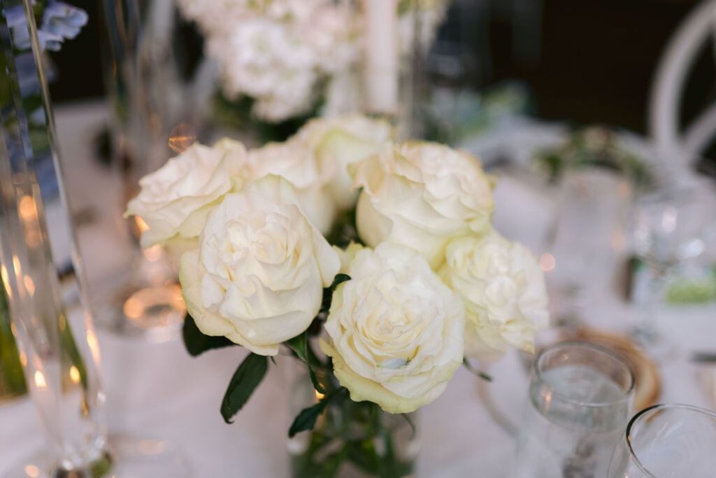 A close-up of a bouquet of white roses in a glass vase, surrounded by glassware and soft, out-of-focus table decor in the background.