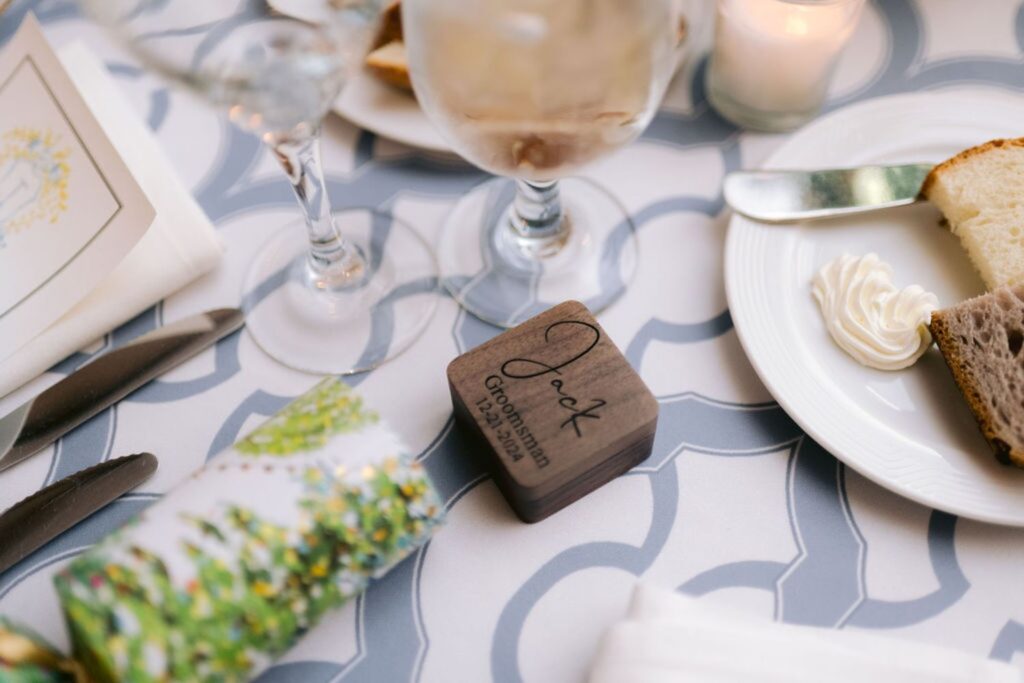 A close-up of a decorated table setting with a wooden box engraved with Jack, Groomsman, 09.29.18, a plate with bread and butter, cutlery, a glass of water, and a candle on a patterned tablecloth.