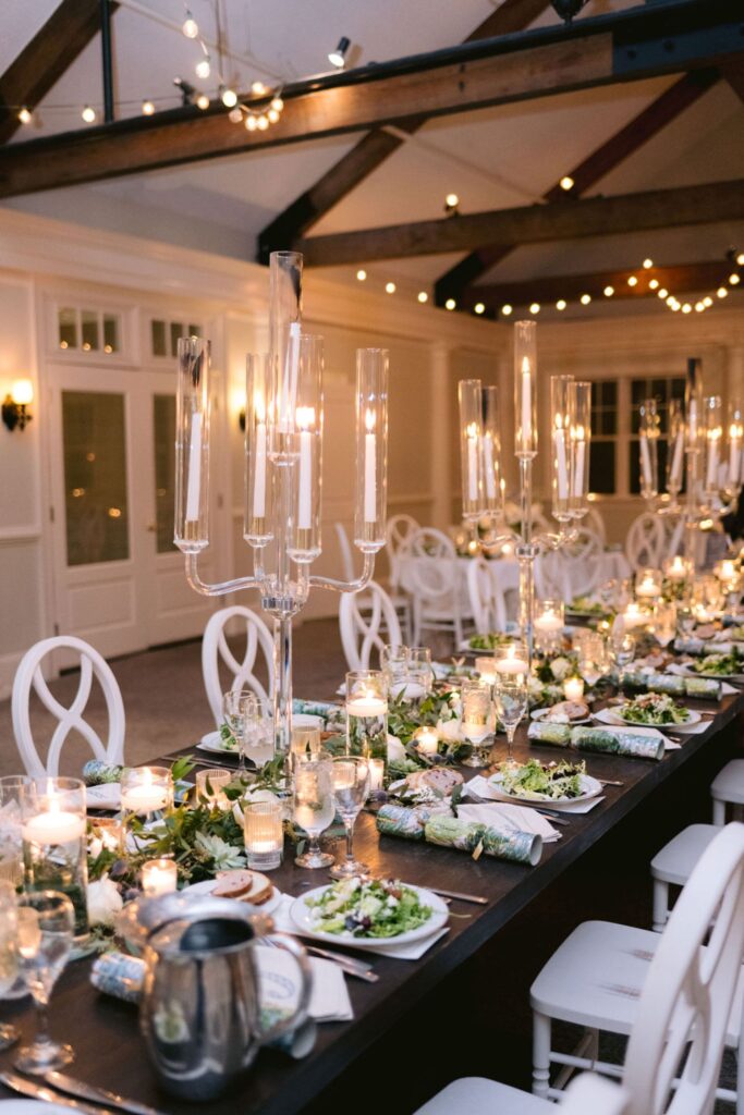 A long, elegant dining table set for a formal event with white chairs, tall glass candle holders, plates of salad, and glowing candles. String lights hang from the ceiling, creating a warm, inviting atmosphere.