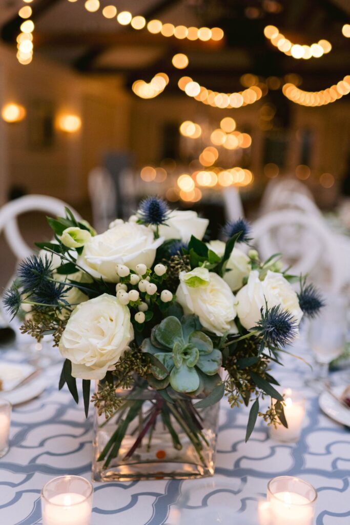 A floral centerpiece with white roses, green succulents, and blue thistles sits in a square glass vase on a patterned tablecloth, surrounded by glowing candles, with warm string lights blurred in the background.