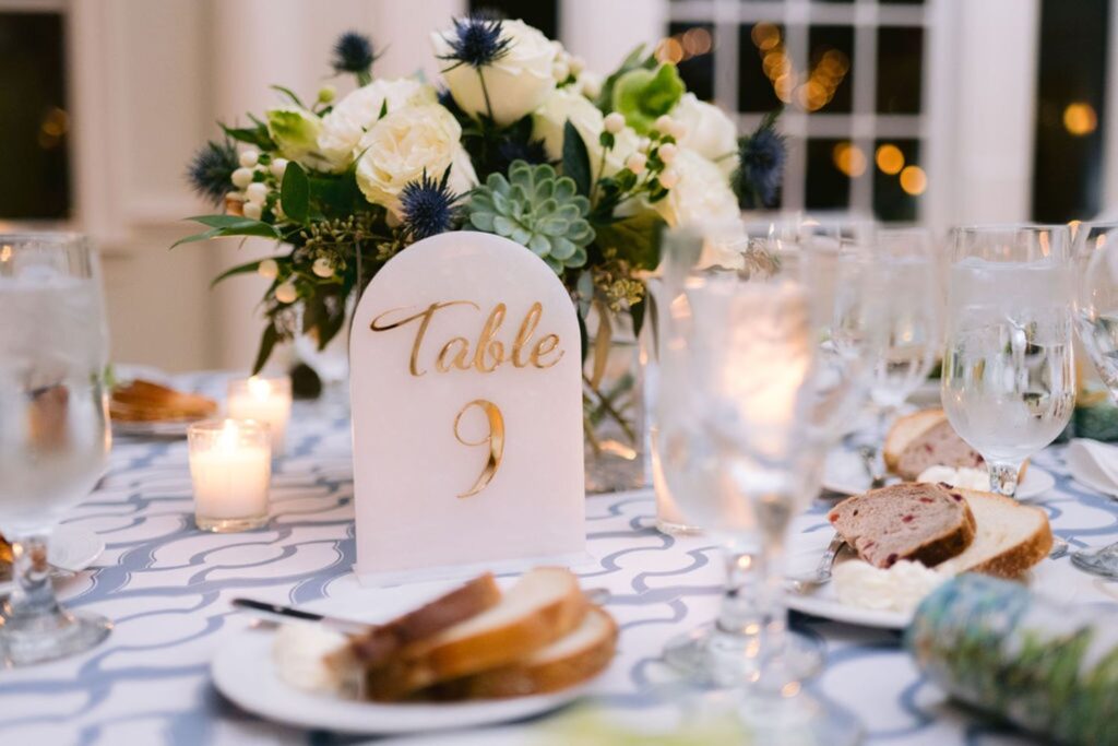 A wedding table with a Table 9 sign, floral centerpiece, lit candles, glasses of water, and plates with bread and butter on a patterned tablecloth.