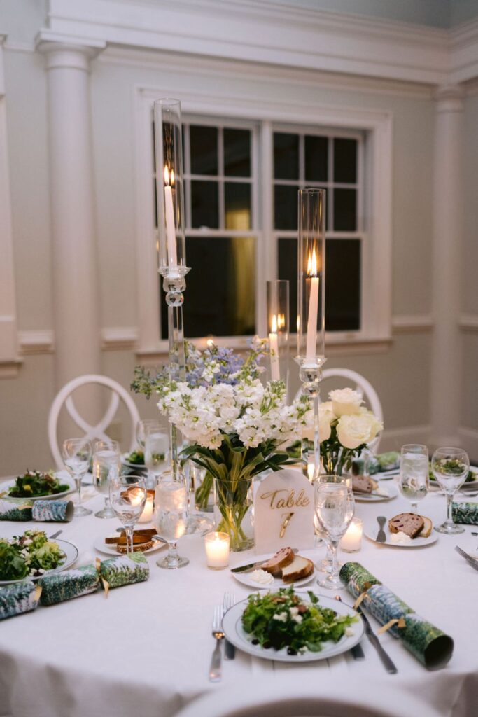 A round table set for a formal event with white chairs, plates of salad, bread, and wrapped favors. The centerpiece features white flowers and tall lit candles. A Table 8 sign is displayed among the decorations.