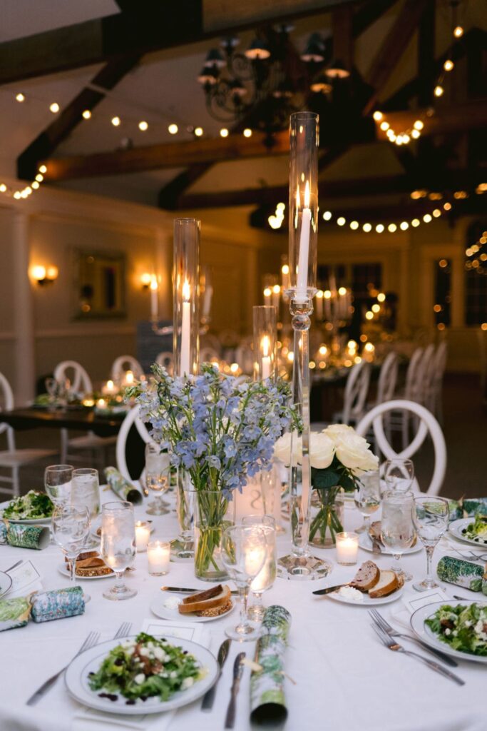 A decorated dining table set for an event with salads, candles, tall glass vases with flowers, and string lights overhead creating a warm, festive ambiance in a spacious room.