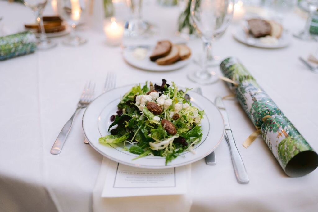 A plate of mixed green salad with crumbled cheese and nuts sits on a white tablecloth, surrounded by cutlery, a Christmas cracker, glasses, a menu, candles, and slices of bread in the background.
