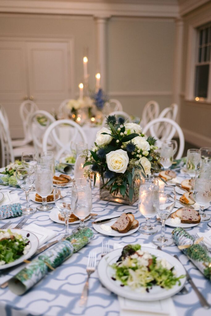 A formal dining table set with salads, bread, glasses of water, and elegant tableware. The centerpiece is a floral arrangement with white roses and greenery. The background shows more decorated tables and lit candles.
