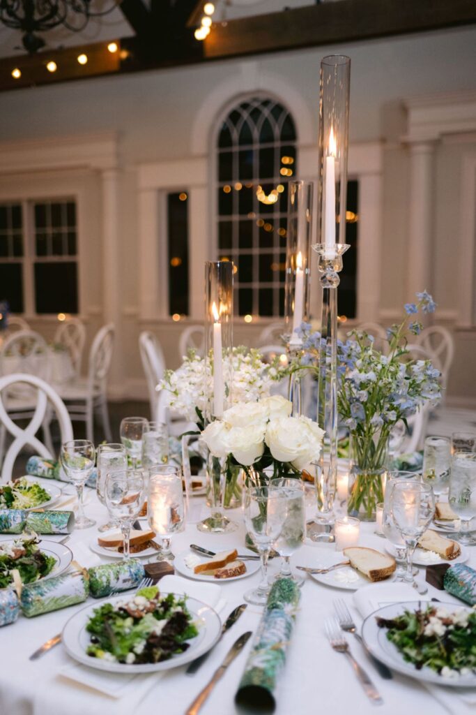 A beautifully set round dining table with plates of salad, bread, water glasses, and floral-patterned crackers. Tall glass candle holders, white roses, and blue flowers serve as the centerpiece in an elegant, softly lit room.