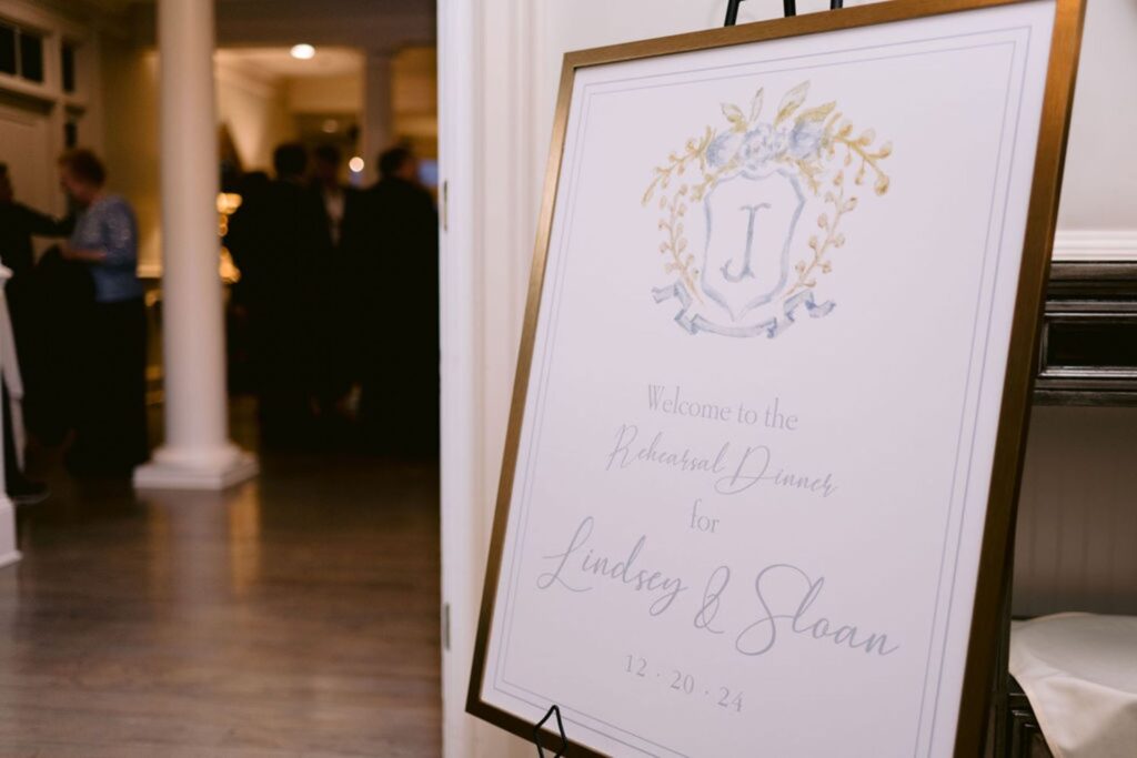 A decorative wedding welcome sign on an easel reads: Welcome to the Rehearsal Dinner for Lindsey & Sloan 12-20-24, with a floral crest and letter J at the top. Blurred guests are visible in the background.