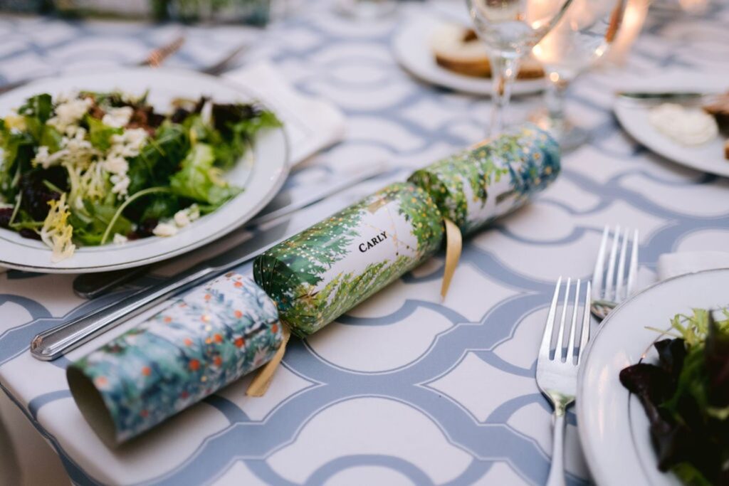 A festive Christmas cracker with a name tag reading CARLY is placed on a patterned tablecloth beside a plate of salad, silverware, glasses, and a lit candle.