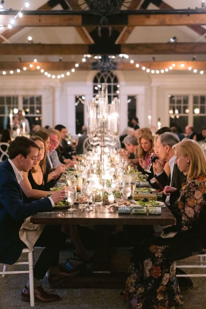 A group of people sit around a long, elegantly set dinner table under string lights in a warmly lit indoor space, enjoying a meal together with candles and decorative glass candelabras as the centerpiece.