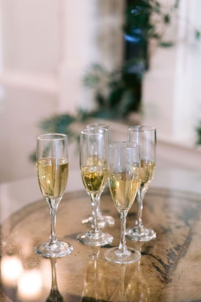 Four champagne flutes filled with sparkling wine are arranged on a round glass table, with a blurred background of greenery and a light-colored interior.