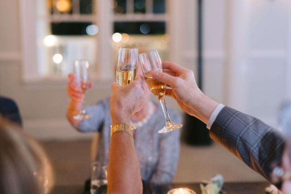 Three people raise champagne glasses in a toast at an indoor evening event, with blurred lights and windows in the background, and one person wearing a plaid jacket sleeve in the foreground.