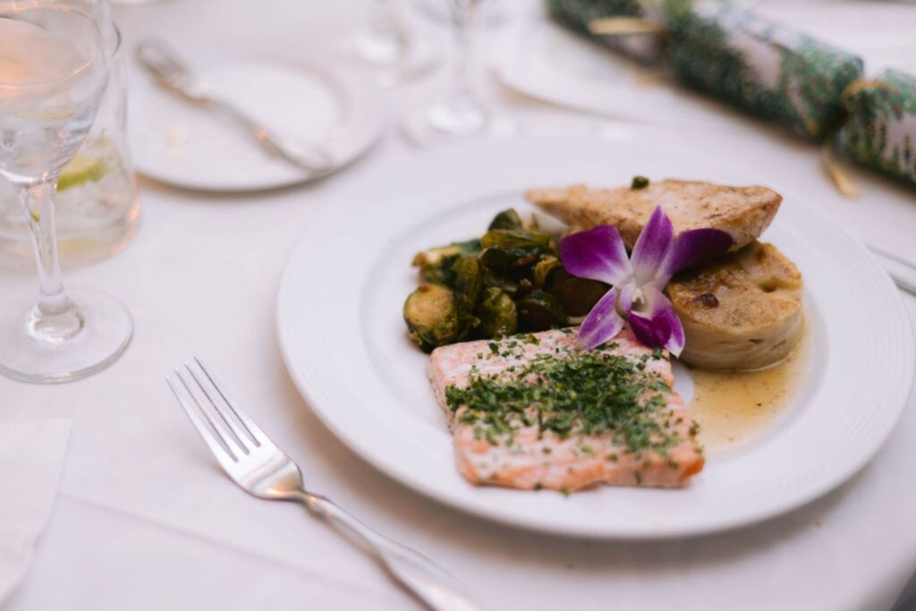 A plated meal featuring a slice of herb-crusted salmon, roasted Brussels sprouts, a piece of bread, and a small serving of gratin, garnished with a purple orchid on a white tablecloth.