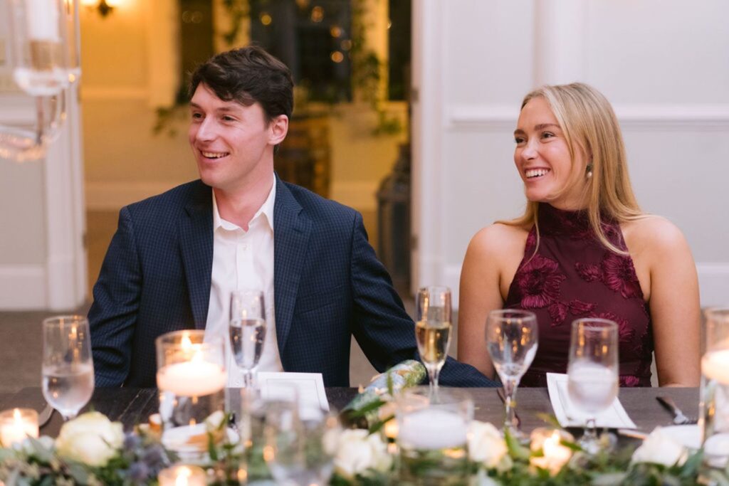 A man in a navy suit and a woman in a burgundy dress sit smiling at a decorated table with candles, glasses, and greenery, in a warmly lit indoor setting.