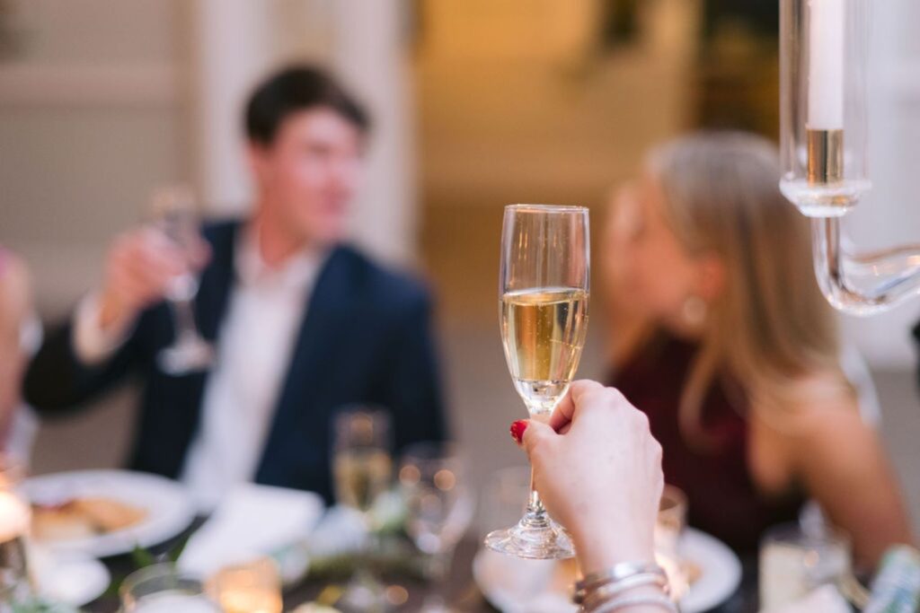 A close-up of a hand holding a glass of champagne in a toast, with two people sitting at a table in the background, enjoying a meal at a festive or celebratory event.