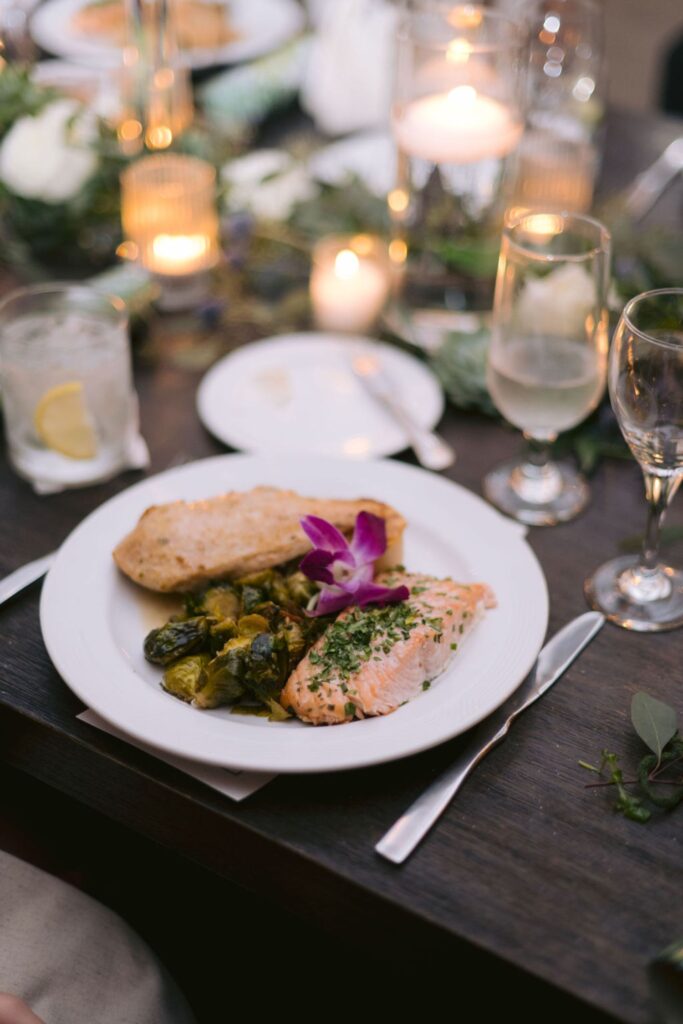 A dinner plate with baked chicken breast, roasted vegetables, and a fillet of salmon garnished with herbs and a purple flower. The table is set with candles, glasses, and greenery in the background.