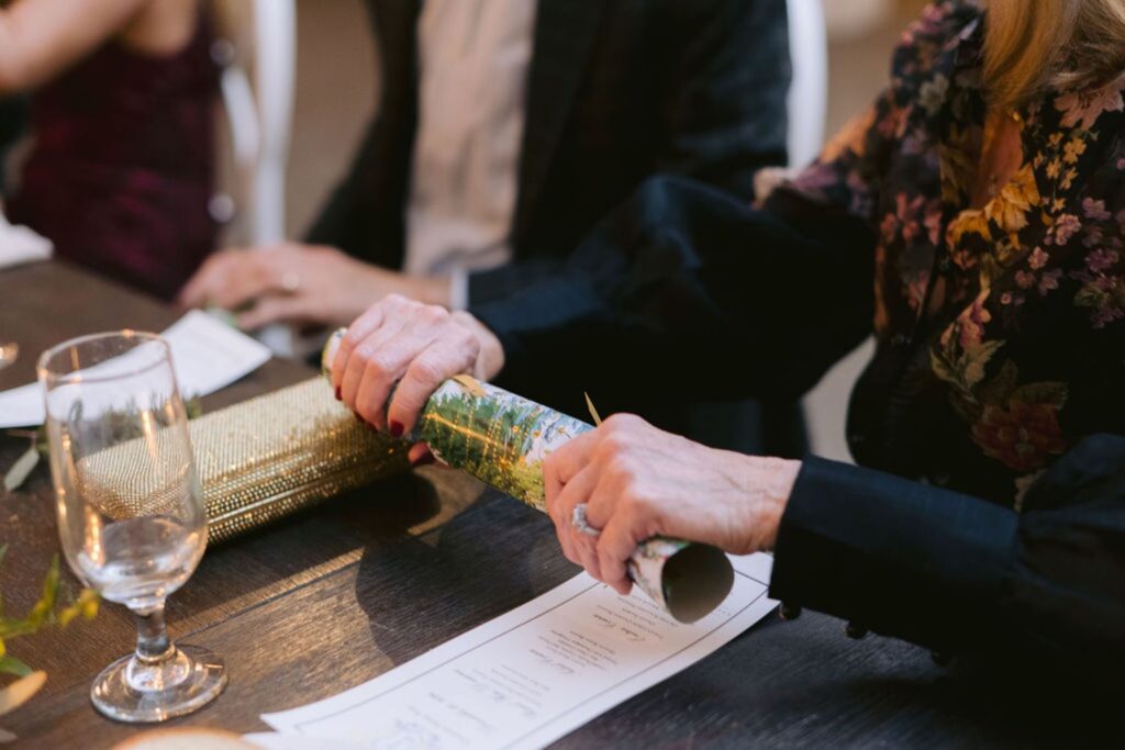 A person in a floral-patterned outfit pulls apart a festive cracker at a table set with menus, a gold clutch purse, a glass, and candles. Another person sits nearby in a dark suit.