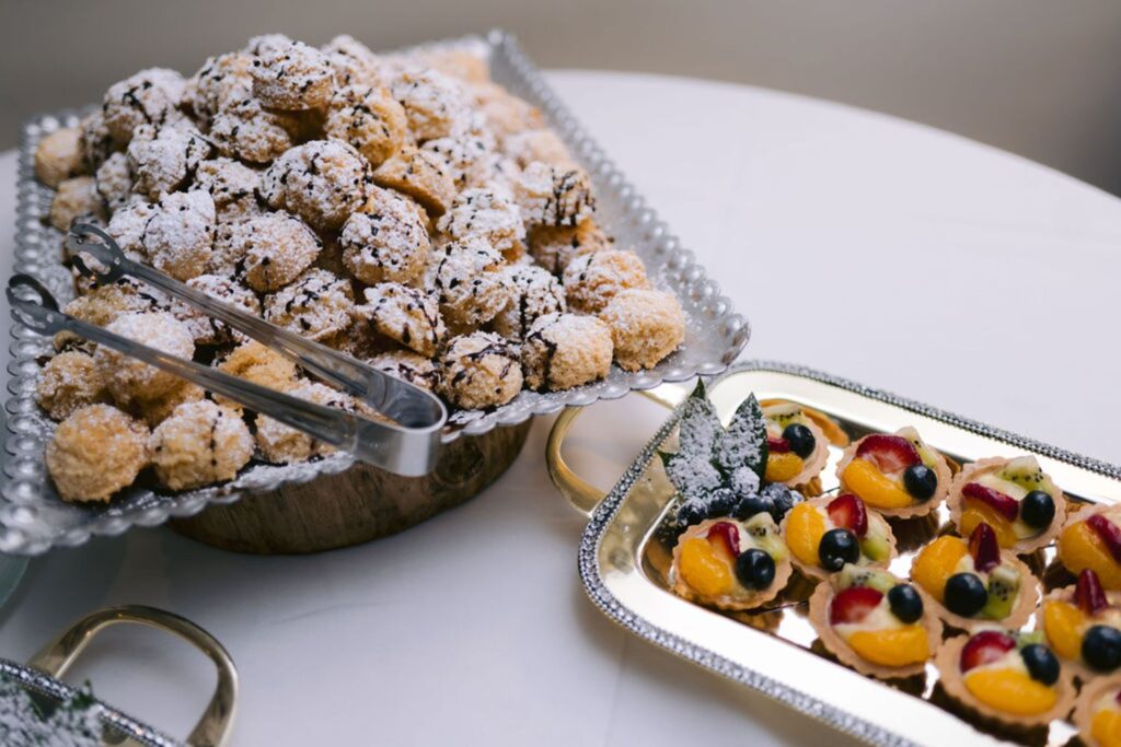 A tray of powdered sugar-coated pastries with tongs beside a platter of fruit-topped mini tarts, displayed on a white tablecloth.