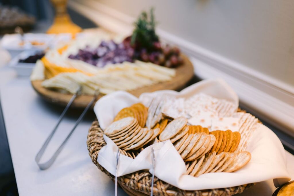 A close-up of a basket filled with assorted crackers on a table, with tongs resting on the side. In the background, a platter of sliced cheese and grapes is slightly out of focus.