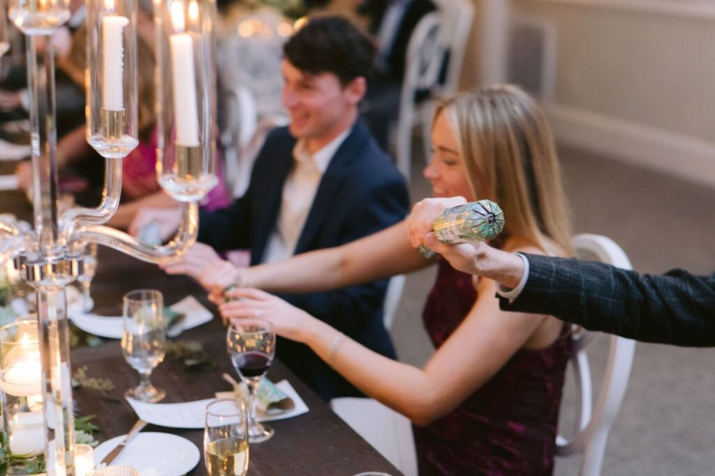 A woman in a burgundy dress and a man in a suit sit at a candlelit dinner table, holding a Christmas cracker together. Other seated guests and elegant table settings are visible in the warmly lit scene.