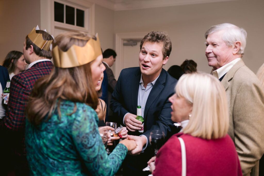 A group of people chatting at a party, some wearing gold paper crowns. They are dressed in semi-formal attire, holding drinks, and appear to be engaged in lively conversation in an indoor setting.
