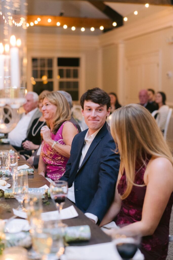 A young man in a suit smiles at a woman sitting beside him at a warmly lit, elegant dinner event. Other people sit around the table, chatting and enjoying the meal. Candles and string lights add a festive ambiance.