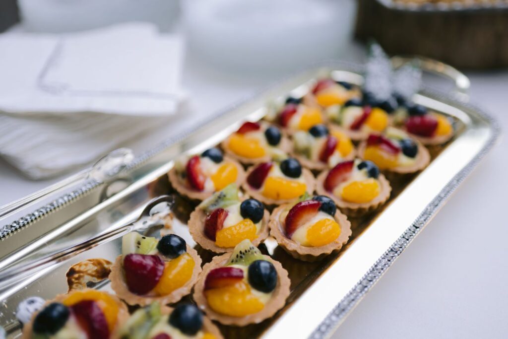 A silver tray holds mini fruit tarts topped with blueberries, strawberries, kiwi, and mandarin slices. Tongs rest on the tray, and napkins are stacked in the background.
