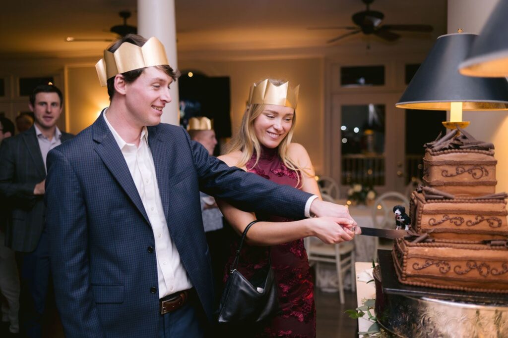 A smiling couple wearing gold paper crowns cut a multi-tiered chocolate cake together at a festive indoor gathering, with guests and soft lighting in the background.