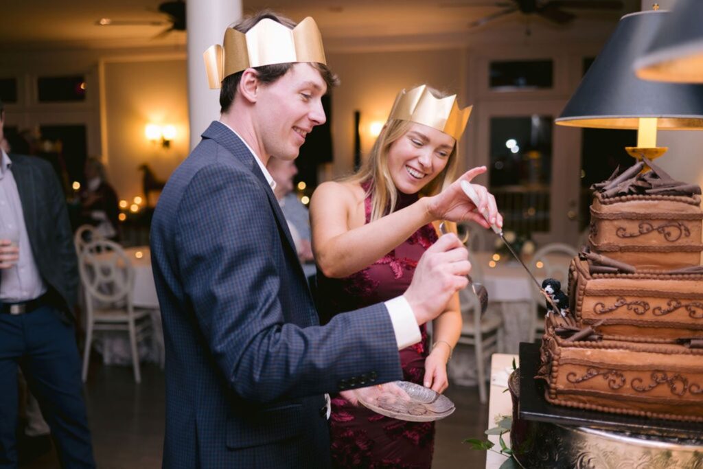 A man and woman wearing gold paper crowns smile as they cut a multi-tiered chocolate cake together at a festive indoor event. The woman holds the knife while the man holds a plate.
