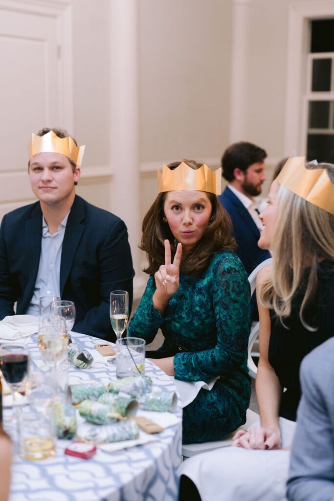 A woman wearing a gold paper crown flashes a peace sign and smiles at the camera while sitting at a table with others during a festive celebration. People around her also wear crowns and appear to be enjoying themselves.