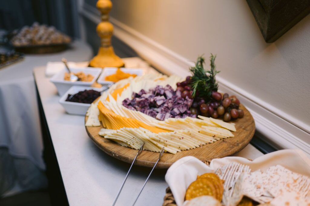 A wooden platter with assorted sliced cheeses, cubed meat, grapes, and herbs, displayed on a white tablecloth. Nearby are baskets of crackers and small bowls of snacks.