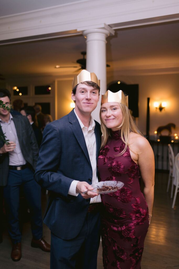 A man in a suit and a woman in a burgundy dress, both wearing gold paper crowns, smile together at a party. The man holds a plate of dessert. People and warm lighting are visible in the background.