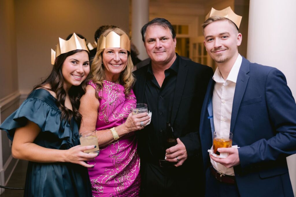 Four adults wearing gold paper crowns smile at the camera while holding drinks at an indoor event. The women wear an off-shoulder green dress and a pink dress; the men wear a black shirt and a navy suit.