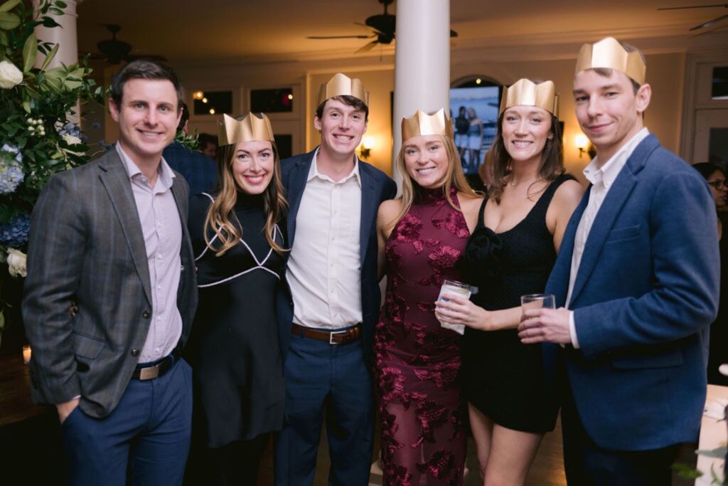 Six young adults wearing gold paper crowns pose together, smiling at a festive indoor party. Three are dressed in jackets, and three in dresses, with two holding drinks. The atmosphere appears cheerful and celebratory.