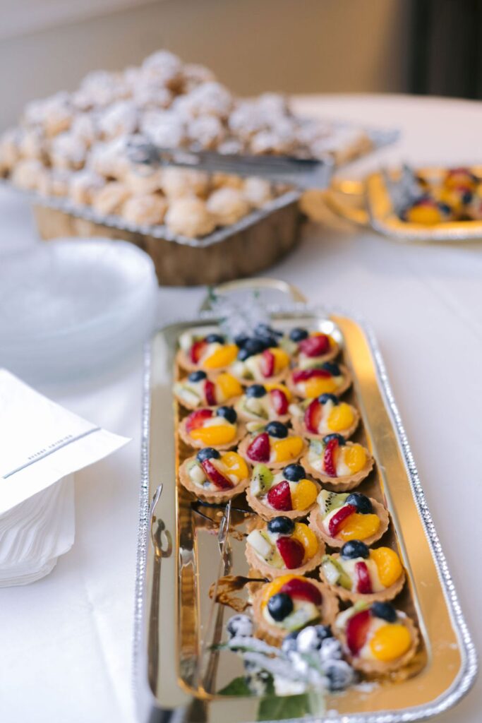 A silver tray with colorful mini fruit tarts topped with berries and kiwi sits on a table, with a platter of powdered sugar pastries and stacks of plates in the background.