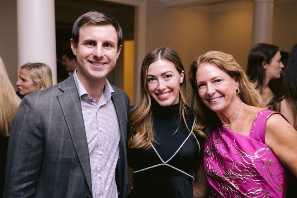 Three people, two women and one man, smiling and posing together at a formal indoor event. The woman on the right is wearing a bright pink dress with a decorative pattern. Other people are visible in the background.