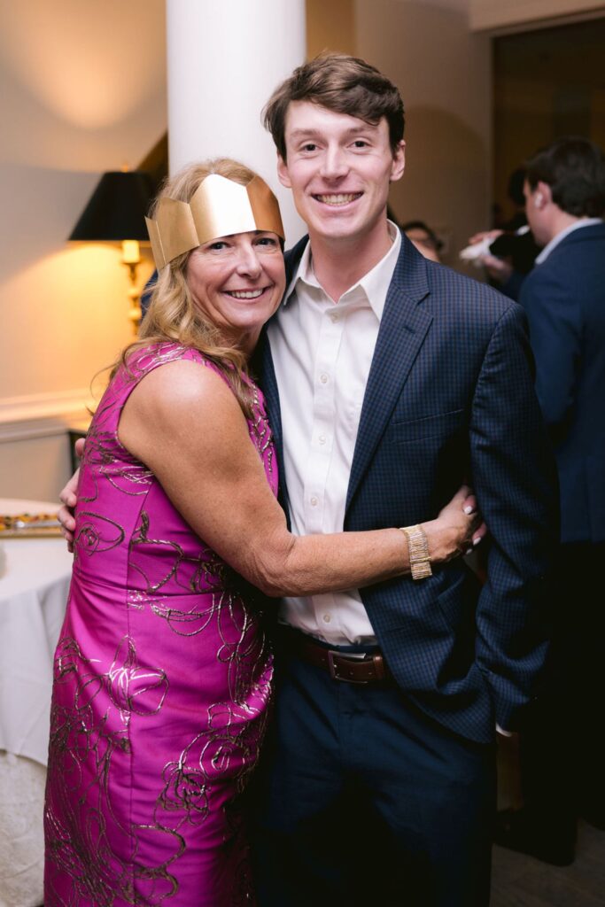 A woman in a shiny pink dress and gold paper crown hugs a smiling young man in a dark suit at an indoor event or party. Other people and tables are visible in the background.