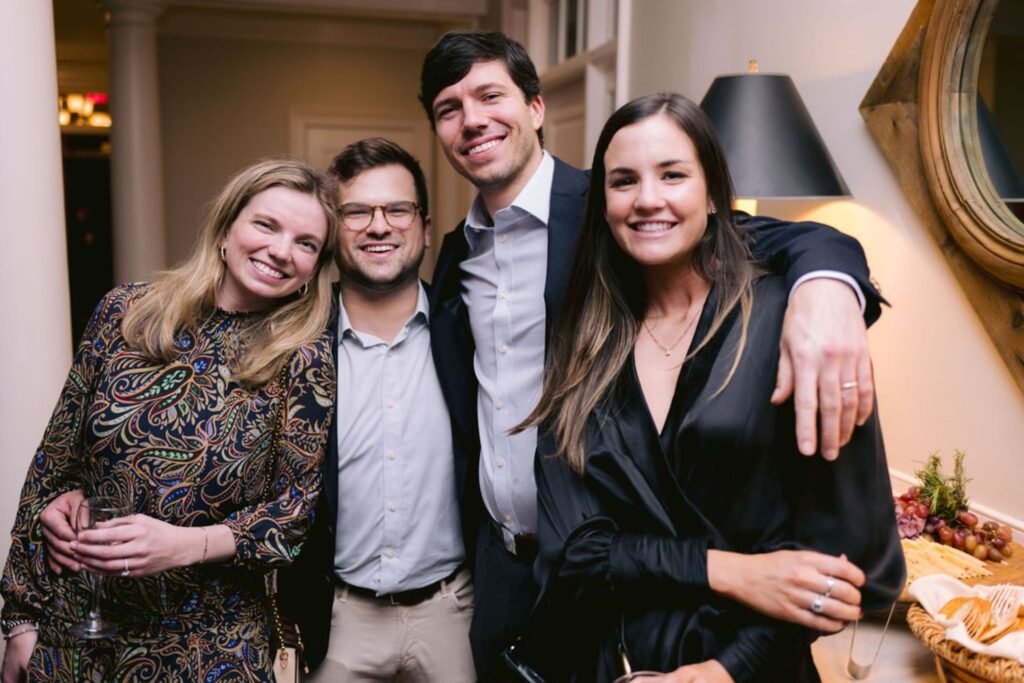 Four adults dressed in semi-formal attire stand closely together, smiling at the camera in an indoor setting. A round mirror and wine glasses are visible in the background.