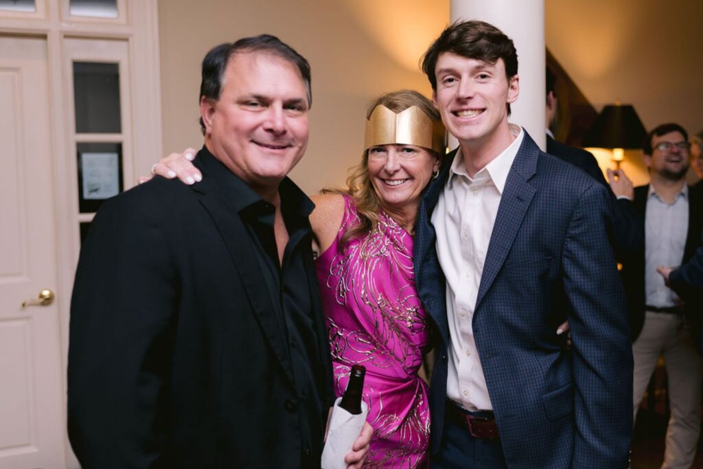 Three people dressed in formal attire smile and pose together at a party; the woman in the center wears a pink dress and a gold headband, while the men on either side wear suits. Other guests are visible in the background.