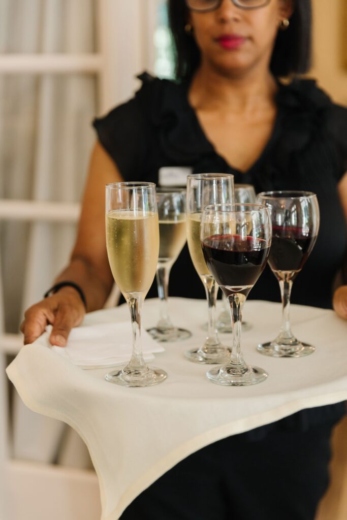 At a Chastain Horse Park rehearsal dinner, a woman holds a tray covered with a white cloth, carrying glasses of red wine and champagne. She wears glasses and a black top. The softly blurred background adds elegant ambiance.