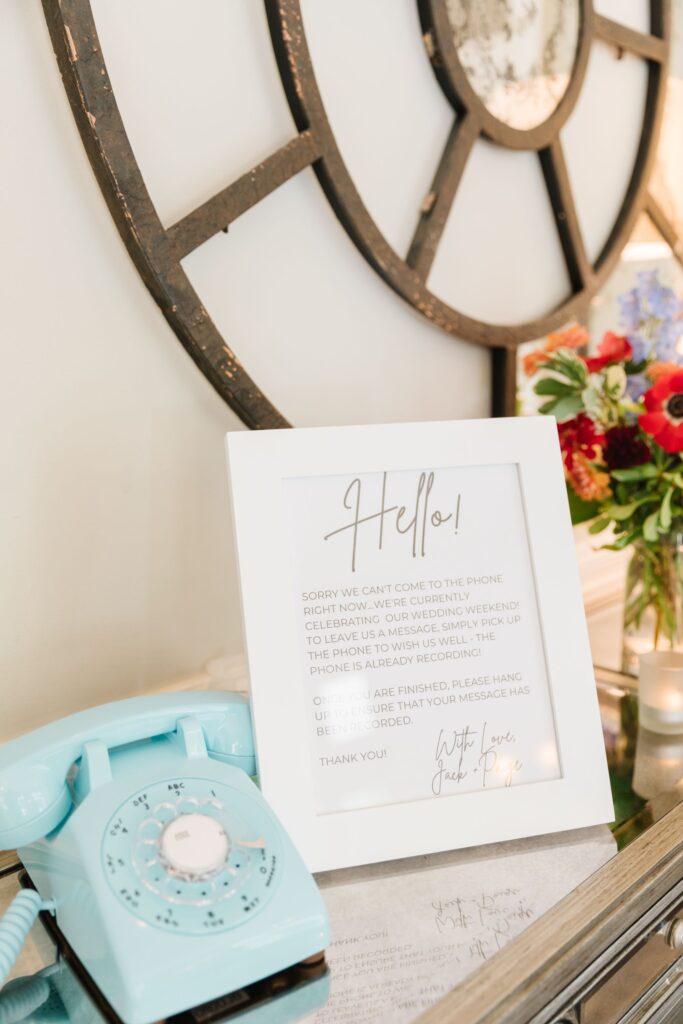 A light blue rotary phone sits on a table next to a framed "Hello!" sign and flowers in a vase. Behind them, a decorative clock hangs on the wall—perfect touches for a Chastain Horse Park rehearsal dinner setting.