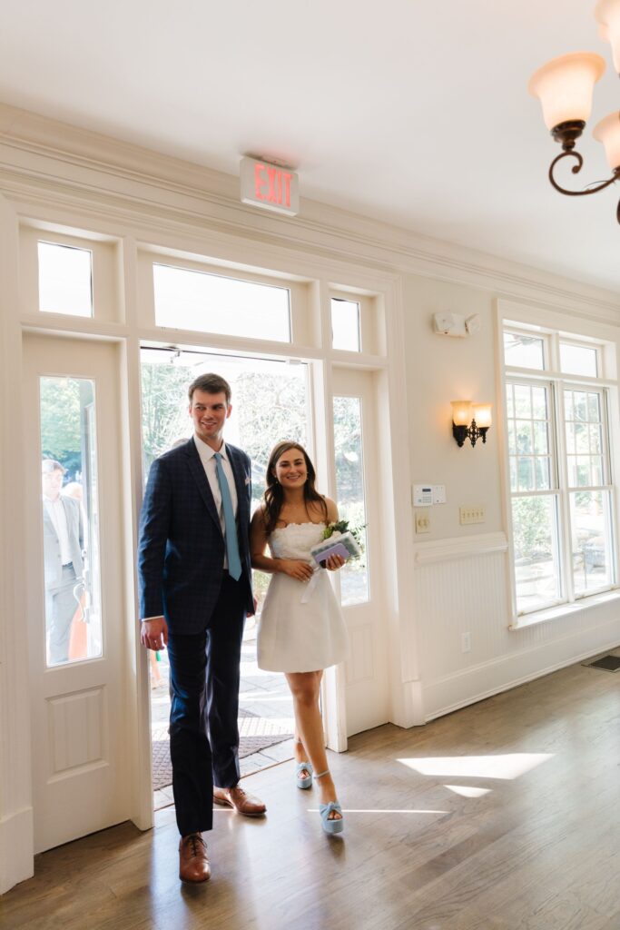 A smiling couple, dressed in formal attire, enters a sunlit room at Chastain Horse Park through glass doors. The woman wears a short white dress and holds flowers; the man wears a blue suit and tie. Warm light streams through the large windows.