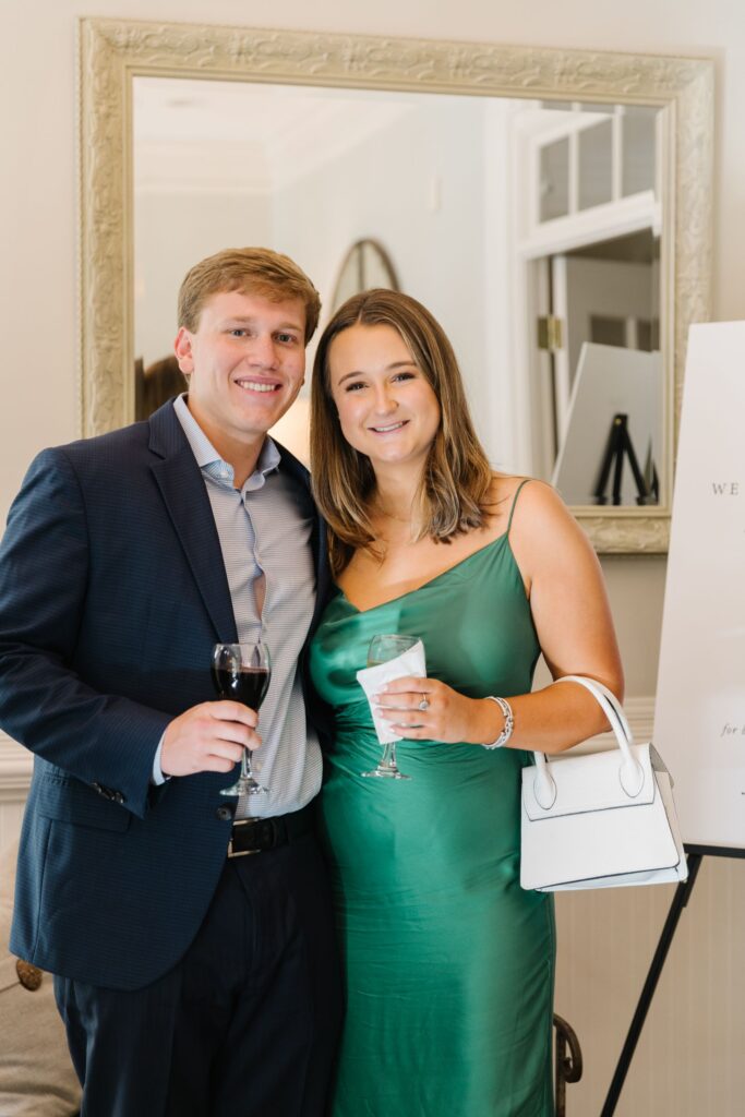 A man in a dark suit and a woman in a green dress stand close together, smiling and holding drinks at a Chastain Horse Park rehearsal dinner. The woman carries a white handbag. They are indoors, with a mirror and white sign in the background.