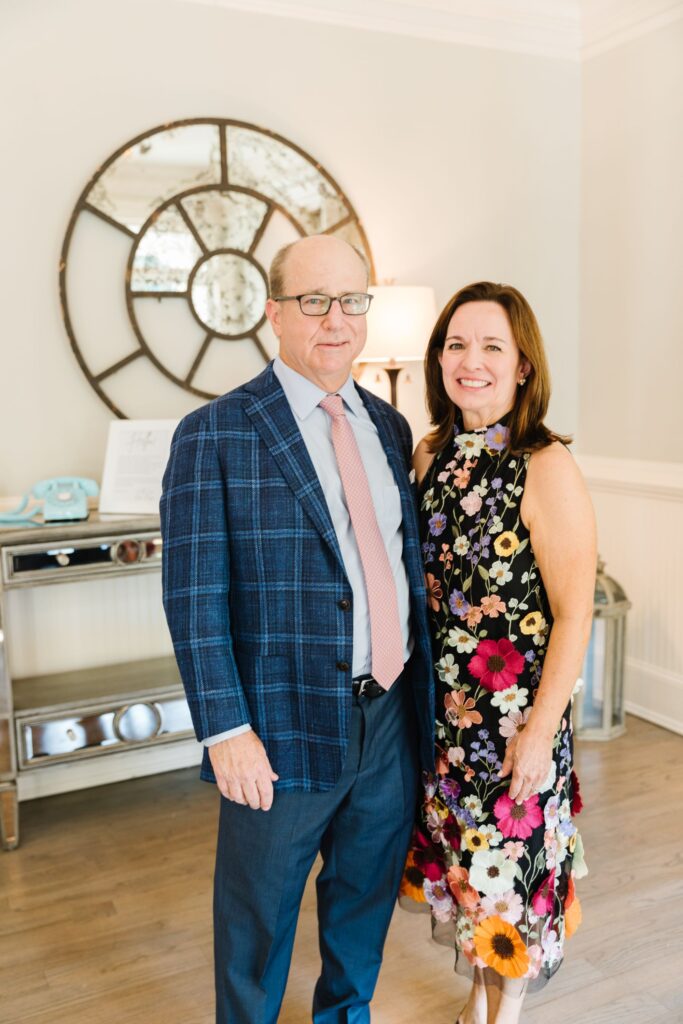 A man in a blue plaid suit and pink tie stands beside a woman in a sleeveless floral dress. Smiling indoors, they celebrate their Chastain Horse Park rehearsal dinner, with a round decorative mirror and console table behind them.