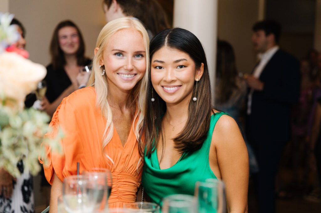 Two women smile and pose together at a Chastain Horse Park rehearsal dinner. One wears an orange dress, the other a green dress. Other guests and glasses are visible in the blurred background, suggesting a festive, indoor gathering.