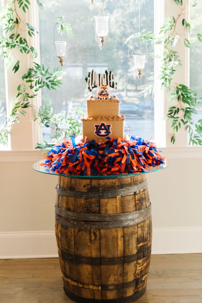 A two-tiered cake with a tiger-striped topper sits on a glass table over a wooden barrel at a Chastain Horse Park rehearsal dinner, surrounded by blue and orange pom-poms, an Auburn University logo, hanging lights, and green vines.
