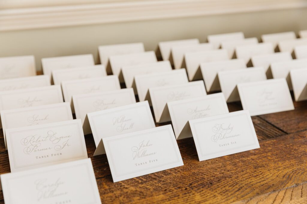 Rows of white, tented place cards with elegant calligraphy are arranged on a wooden surface, each displaying a guest’s name and table number for a formal event or Chastain Horse Park rehearsal dinner.