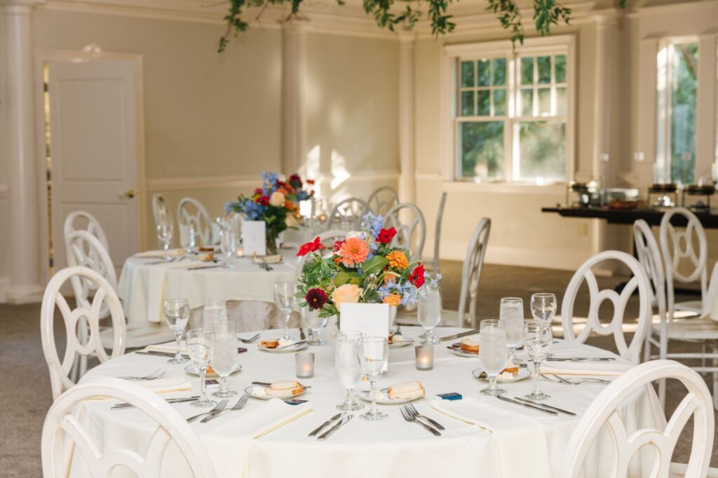 Round tables set for a formal Chastain Horse Park rehearsal dinner, each with white tablecloths, floral centerpieces, glassware, plates, and silverware. White chairs surround the tables in a bright room with windows and greenery overhead.